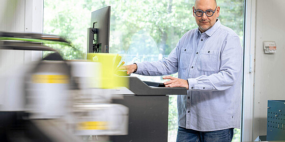 Ein Mitarbeiter bedient konzentriert ein Steuerpult an einer CNC-Schneidemaschine für Kartonagen vor einem großen Fenster.