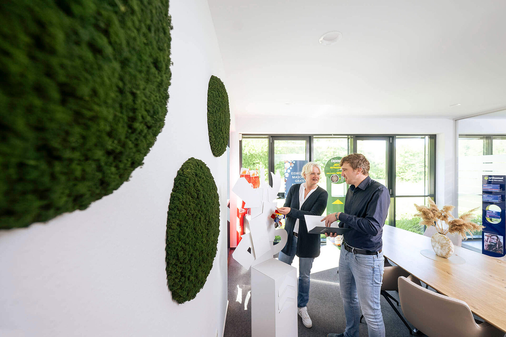 Eine Frau und ein Mann stehen in einem modernen Büro mit Moos-Wanddeko und besprechen eine weiße Skulptur aus Kartonelementen.