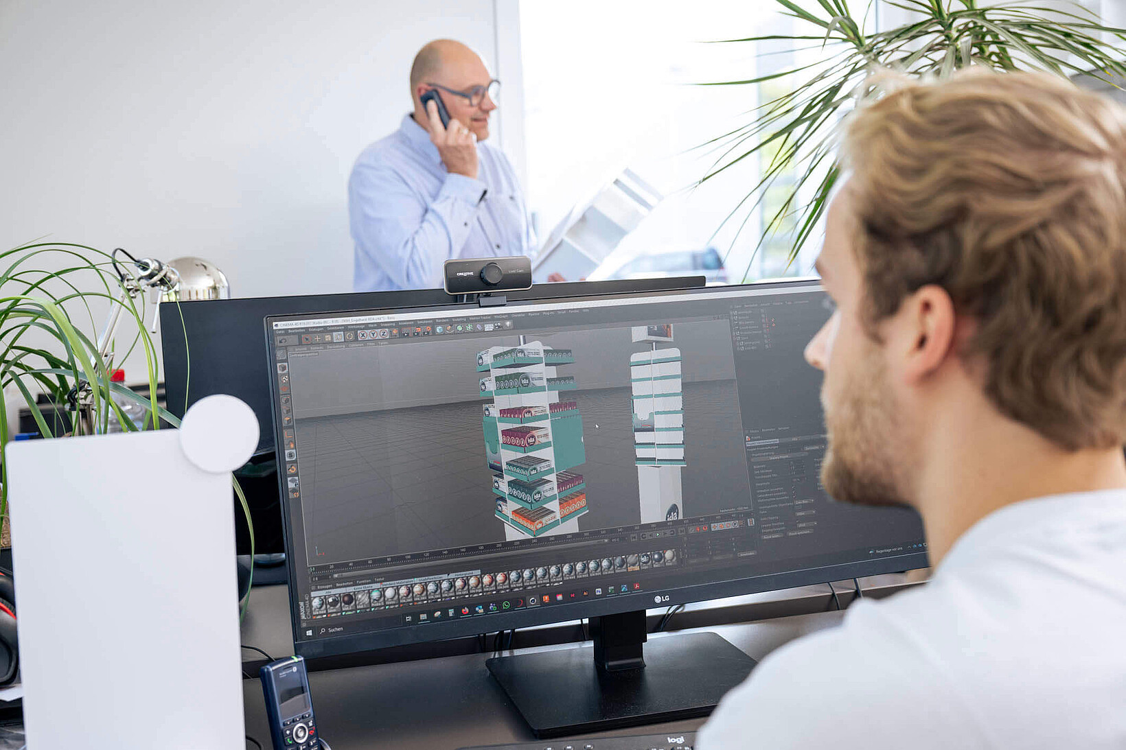 Ein Mitarbeiter arbeitet im Vordergrund an einem großen Monitor an einem 3D-Modell eines Verkaufsdisplays, während im Hintergrund ein Kollege telefoniert.