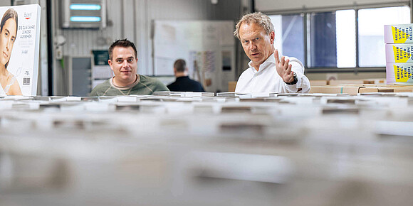 Zwei Mitarbeiter in einer Produktionshalle blicken über eine Reihe von verpackten Produkten hinweg in die Kamera; im Hintergrund ist Werbe- und Displaymaterial zu sehen.