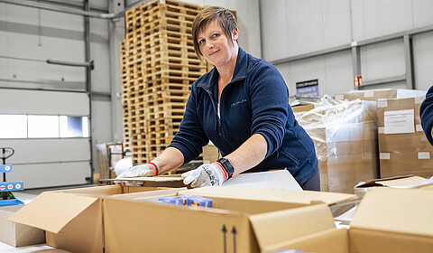 Eine Frau mit kurzen braunen Haaren und Arbeitshandschuhen verpackt Waren in Kartons in einer Lagerhalle.