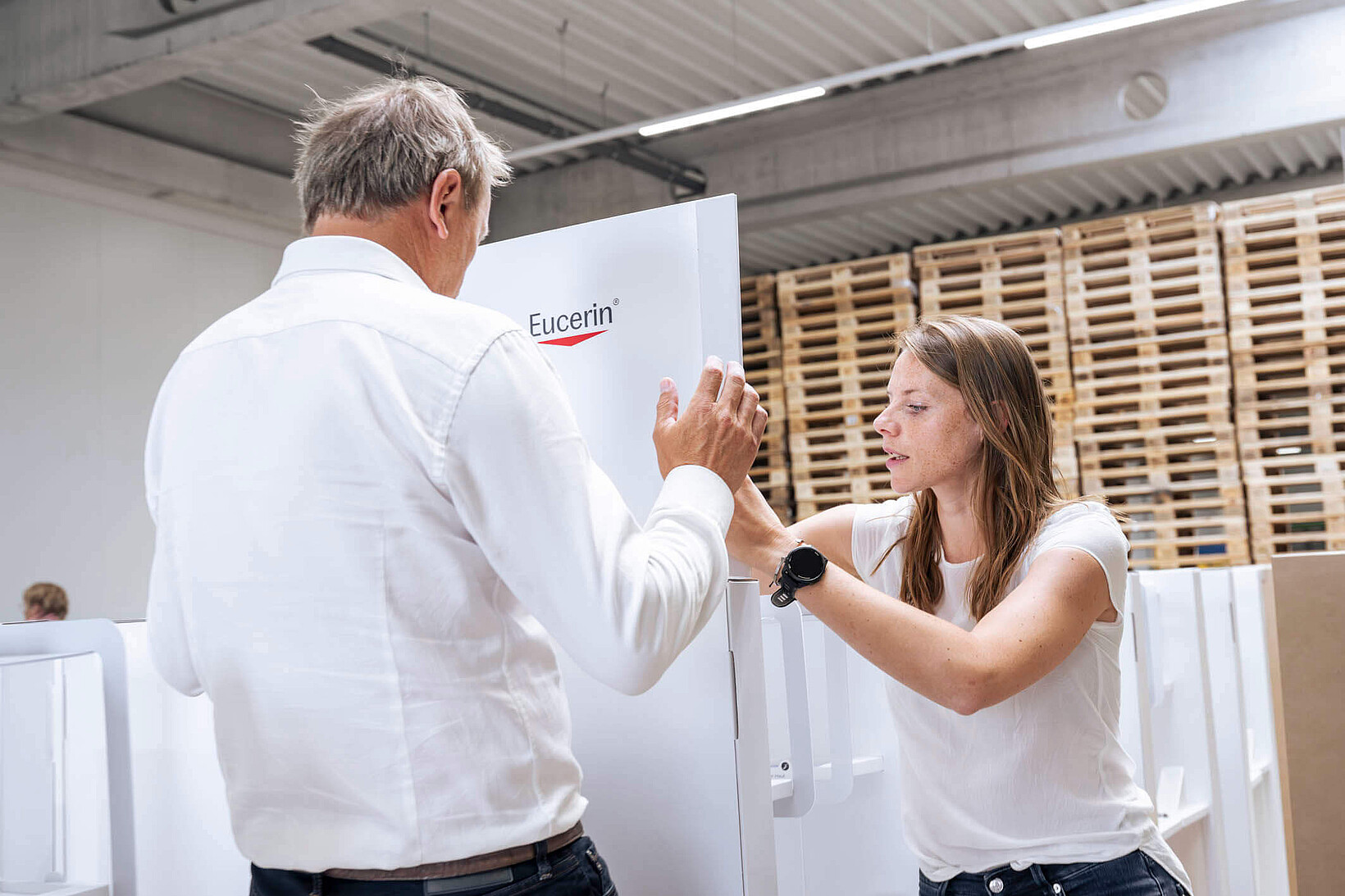 Eine Frau und ein Mann bauen in einer Produktionshalle gemeinsam ein großes, weißes Verkaufsdisplay mit dem Logo „Eucerin“ auf.