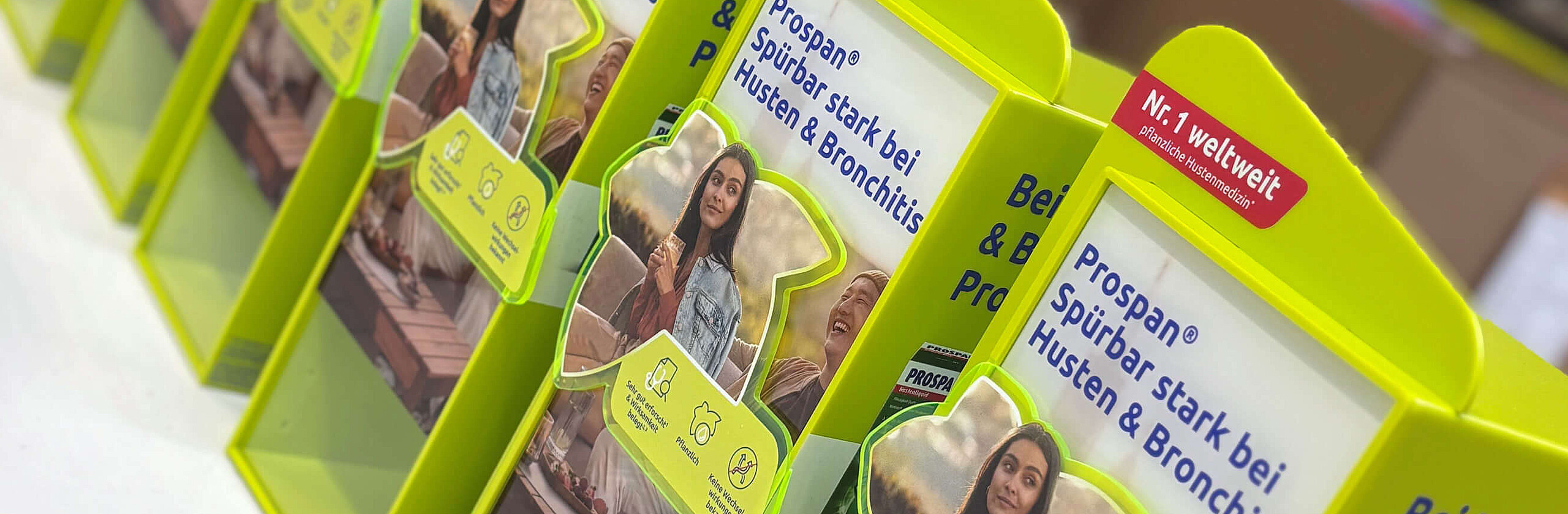 Eine Reihe leuchtend gelb-grüner Prospan-Verkaufsaufsteller mit der Aufschrift „Spürbar stark bei Husten & Bronchitis“ in einer Lagerhalle.