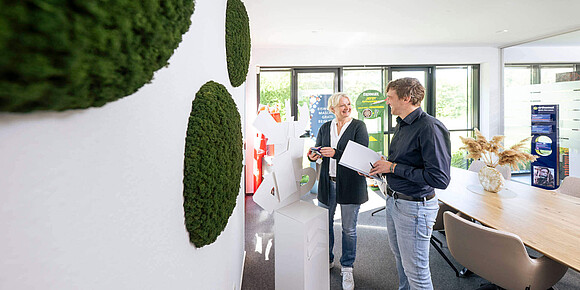 Eine Frau und ein Mann stehen in einem modernen Büro mit Moos-Wanddeko und besprechen eine weiße Skulptur aus Kartonelementen.