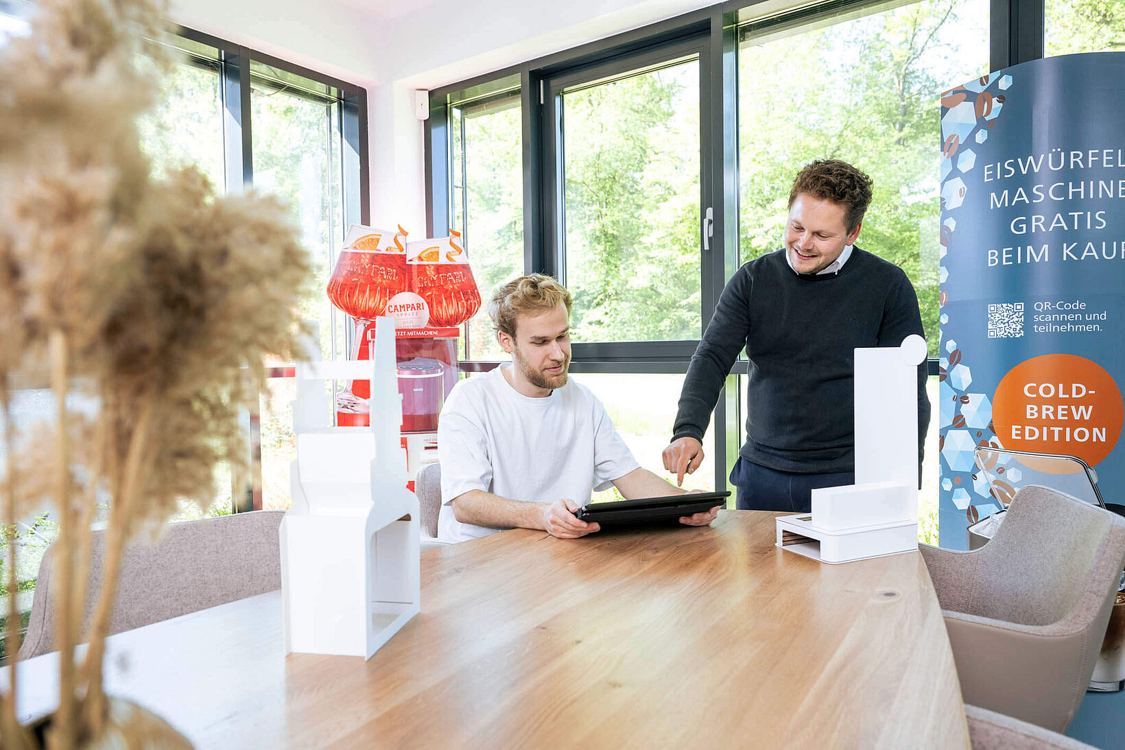 Zwei Mitarbeiter besprechen an einem Tisch ein Projekt auf einem Tablet. Auf dem Tisch stehen weiße Display-Prototypen (Weißmuster), im Hintergrund sind fertige, bedruckte Aufsteller zu sehen.