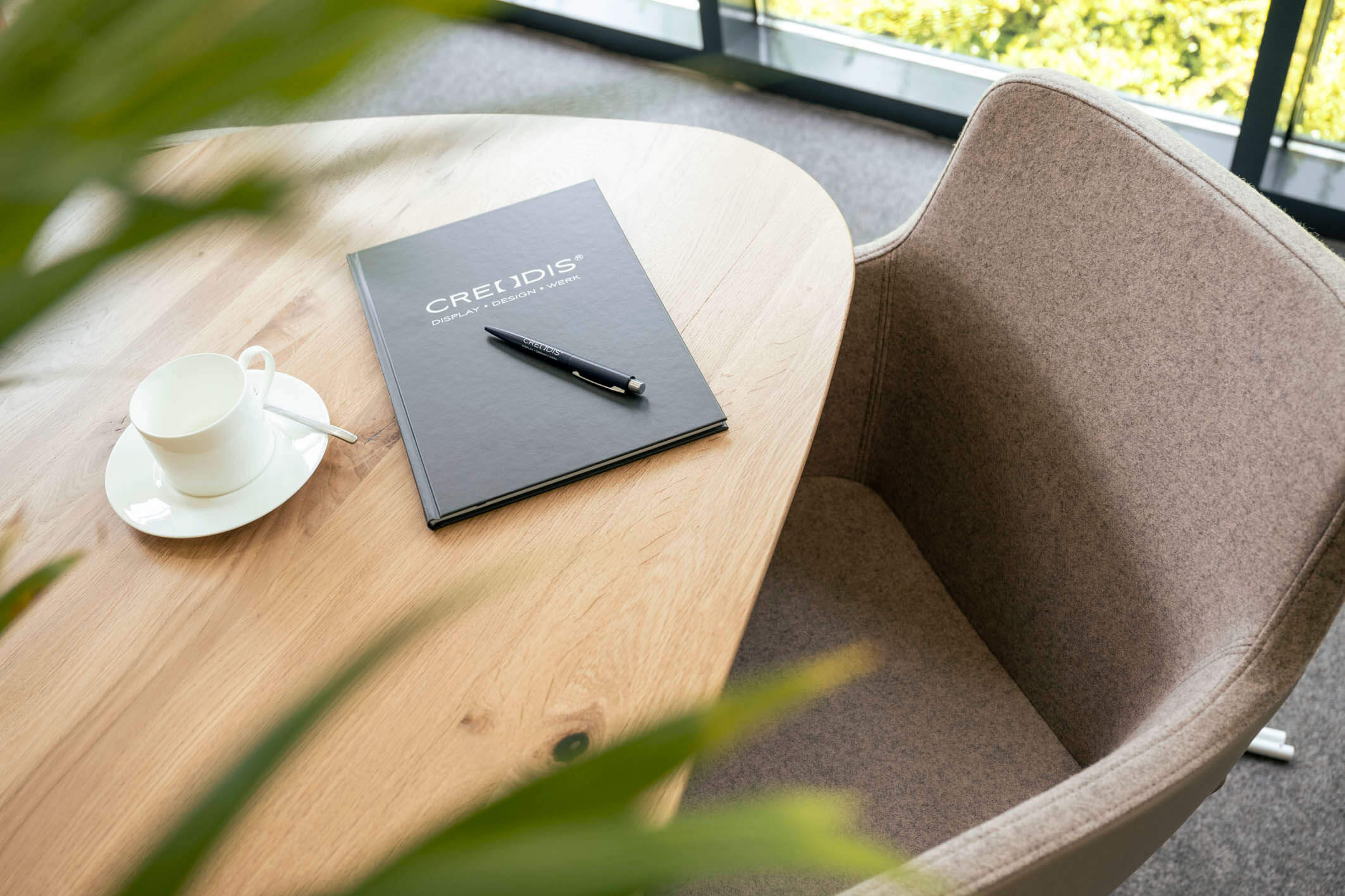 Ein dunkelgraues Notizbuch mit dem silbernen Aufdruck „Creodis Display - Design - Werk“ und ein passender Kugelschreiber liegen auf einem Holztisch neben einer Kaffeetasse.